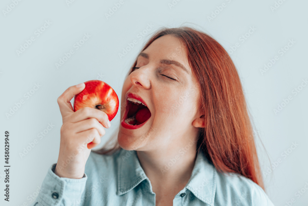 Portrait of a cheerful young woman eating red apple on gray wall background. Healthy nutrition diet. Apple vitamin snack, front view Portrait of a cheerful young woman eating red apple on gray wall background. Healthy nutrition diet. Apple vitamin snack, front view
