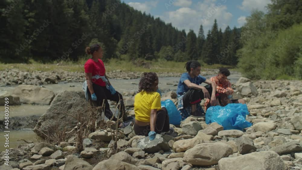 Portrait of exhausted attractive diverse multiethnic people sitting on rocks and relaxing after successful environmental cleanup on mountain riverbank, feeling tired and weary on hot summer day. Portrait of exhausted attractive diverse multiethnic people sitting on rocks and relaxing after successful environmental cleanup on mountain riverbank, feeling tired and weary on hot summer day.
