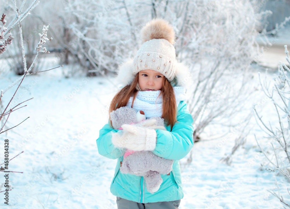 A small, charming girl in winter in a snowy park holds a teddy bear. Background of a winter forest landscape A small, charming girl in winter in a snowy park holds a teddy bear. Background of a winter forest landscape