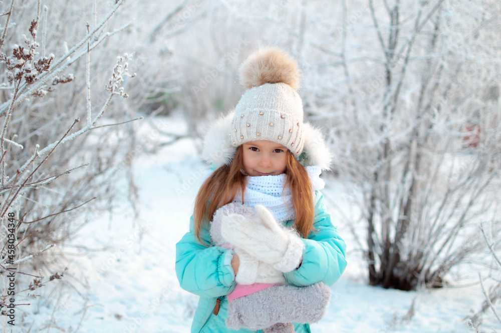 A small, charming girl in winter in a snowy park holds a teddy bear. Background of a winter forest landscape A small, charming girl in winter in a snowy park holds a teddy bear. Background of a winter forest landscape