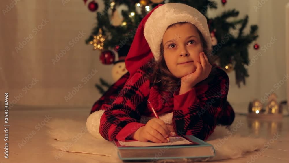 A girl lies on the floor, dreams and writes a letter to Santa Claus, in the background there is a festive Christmas tree. A child in a hat of Santa Claus under the Christmas tree dreams of gifts A girl lies on the floor, dreams and writes a letter to Santa Claus, in the background there is a festive Christmas tree. A child in a hat of Santa Claus under the Christmas tree dreams of gifts