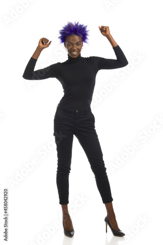 Cheerful African American Woman In Black Clothes And High Heels Is Standing With Arms Raised.