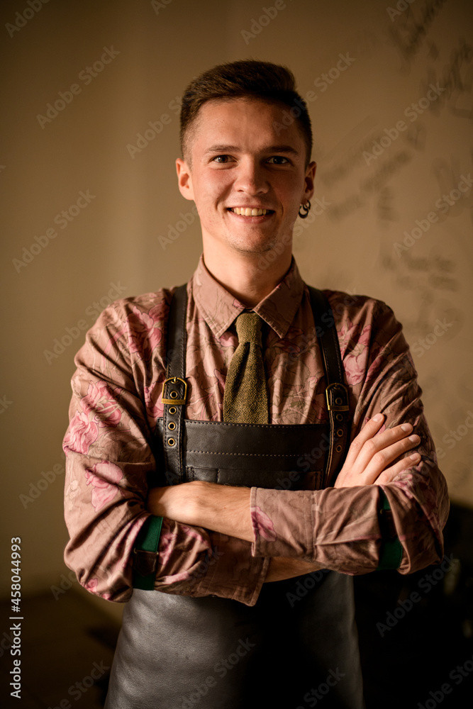 young smiling man bartender in leather apron stands with arms crossed on his chest young smiling man bartender in leather apron stands with arms crossed on his chest