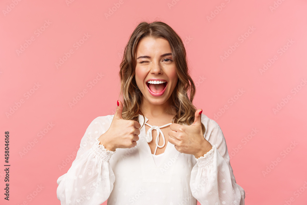 Portrait of sassy good-looking blond girl assure all good, show thumbs-up and wink with classy pleased smile, recommend visit place, shop at online store, standing in white dress pink background - obrazy, fototapety, plakaty 