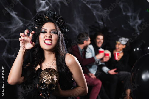 asian woman in vampire halloween costume holding creepy skull and showing scary gesture on black