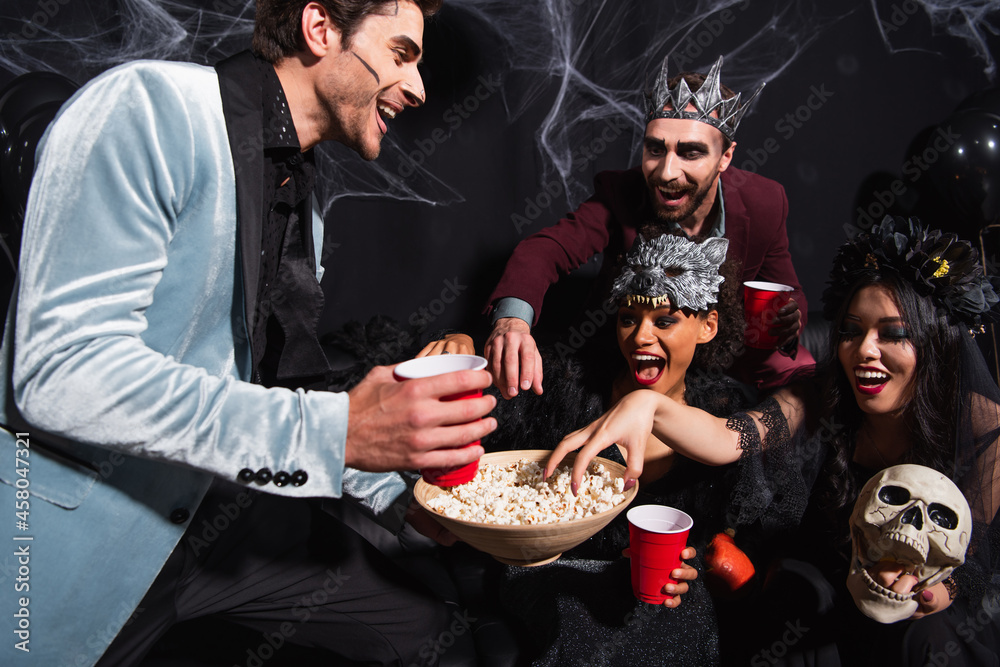 Φωτογραφία thrilled interracial friends eating popcorn during halloween party on black