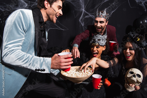 thrilled interracial friends eating popcorn during halloween party on black