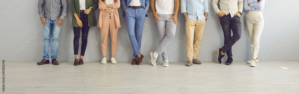 Group of successful confident business people in smart and casual wear standing in studio. Team of employees leaning on grey office wall. Cropped shot of people's legs in classic pants and jeans - obrazy, fototapety, plakaty 