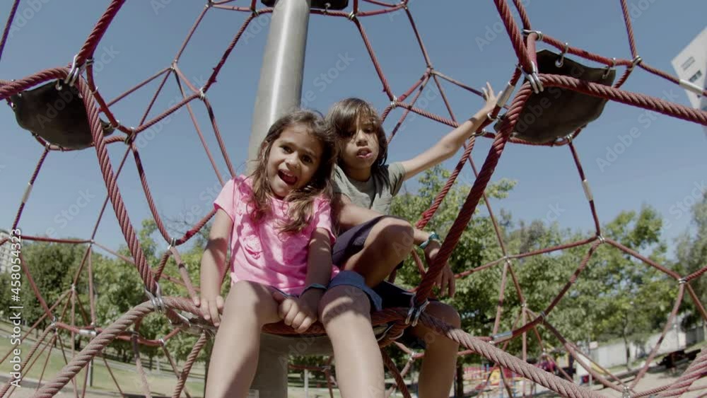 Tired brother and sister sitting on rope attraction. Long shot of Caucasian kids resting after playing on playground, looking at camera and grimacing. Outdoor activity, sport concept Tired brother and sister sitting on rope attraction. Long shot of Caucasian kids resting after playing on playground, looking at camera and grimacing. Outdoor activity, sport concept