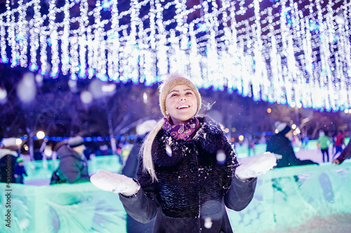 Outdoor photo of young beautiful happy smiling girl holding snow. Background festive Christmas light. Model wearing winter knitted beanie © Parilov
