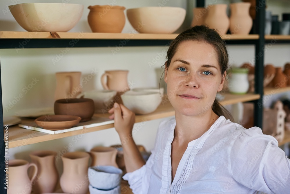 woman artisan ceramist stands on the background of a rack with handmade clay utensils