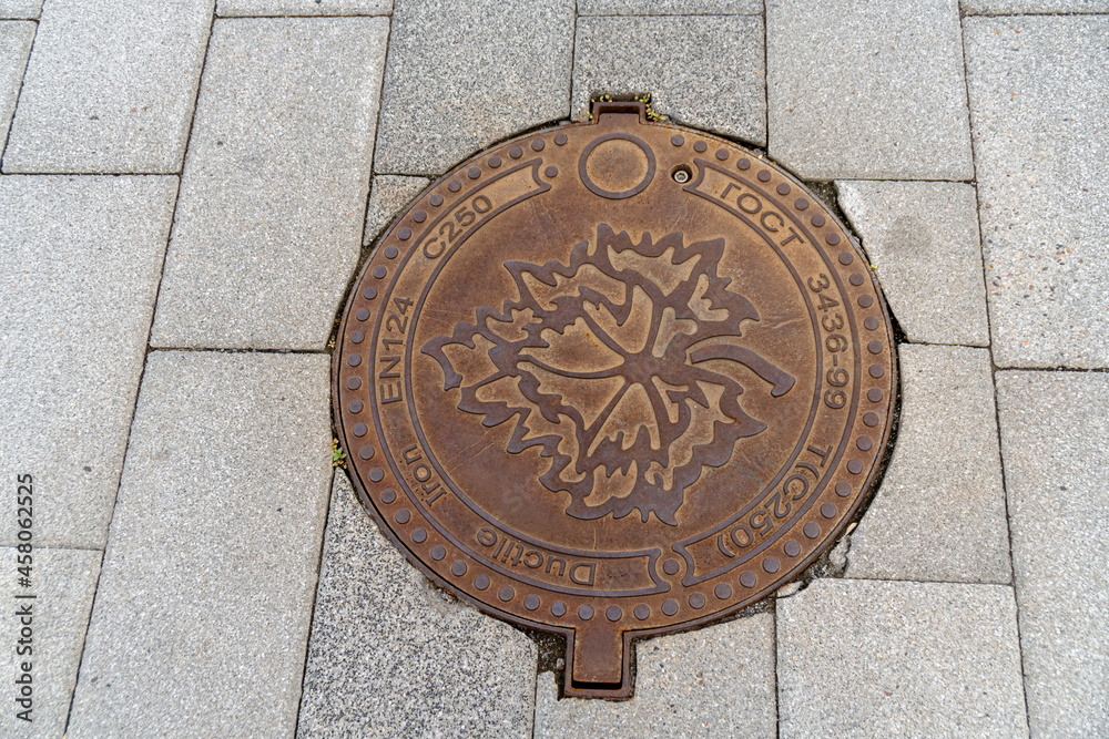 A cast iron manhole cover with embossed maple leaf is positioned on the gray concrete slab sidewalk.