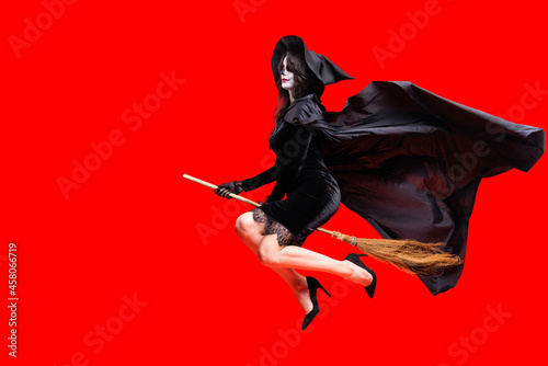 Woman in Halloween costume wear black witch hat, long cloak with creative sugar skull makeup. On red isolated background fly on magic broom. Ð¡oncept Los Muertos poster party or La Calavera Catrina.