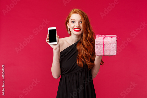 Portrait of excited and amused smiling, wondered redhead woman didnt expact receive gift so fast, showing mobile phone application, online delivery app, hold pink present over red background photo