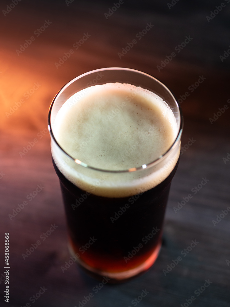 A glass of dark beer on a black wooden table, top view. Orange backlight