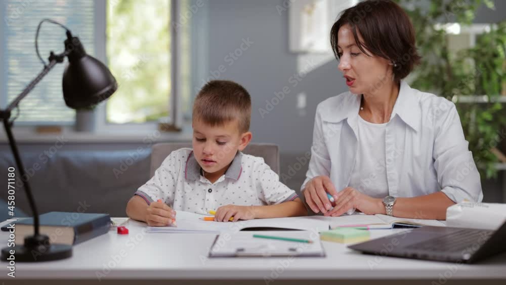 Caring caucasian mother with dark hair helping her son with homework. Schoolboy sitting at table with his mom and writing in copybook. Support and parenthood concept. Caring caucasian mother with dark hair helping her son with homework. Schoolboy sitting at table with his mom and writing in copybook. Support and parenthood concept.