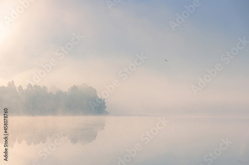 Sunrise over foggy lake. Beautiful dreamy view. Sun is rising up over foggy water and trees with reflections on the river bank.