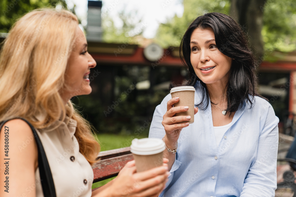 Middle-aged mature caucasian sisters two mature women girlfriends best friends talking spending time together while sitting on the park bench drinking coffee in paper cups. Middle-aged mature caucasian sisters two mature women girlfriends best friends talking spending time together while sitting on the park bench drinking coffee in paper cups.
