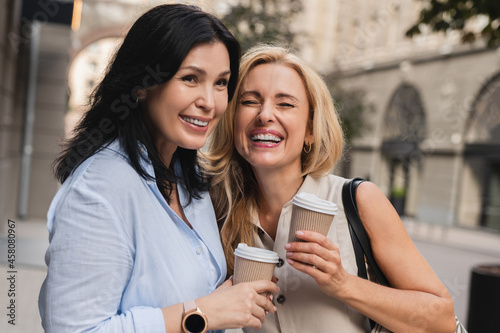 Two cheerful caucasian best friends mature women sisters girlfriends gossiping laughing spending time together while walking on the streets and drinking hot beverage from paper cups outdoors.