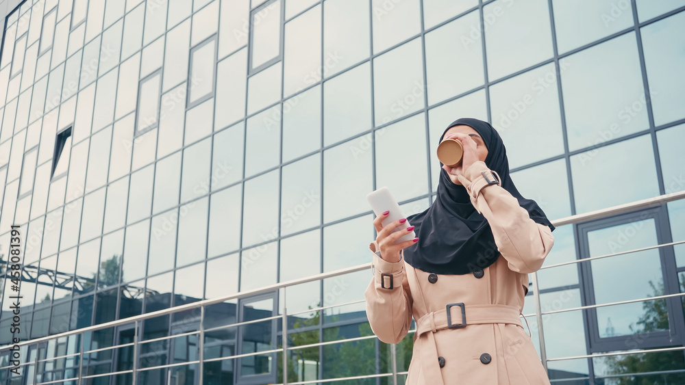 Young muslim woman in hijab holding smartphone and drinking coffee to go near building