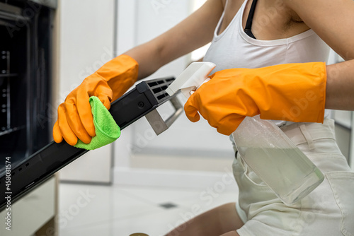 woman in gloves and an apron cleaning oven with rag and spray bottle in kitchen