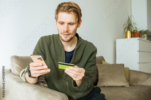 Online shopping from home. Happy man doing internet ecommerce at home. Man working on smart phone. Male using internet banking and making payment. Guy holds smart phone and credit card.