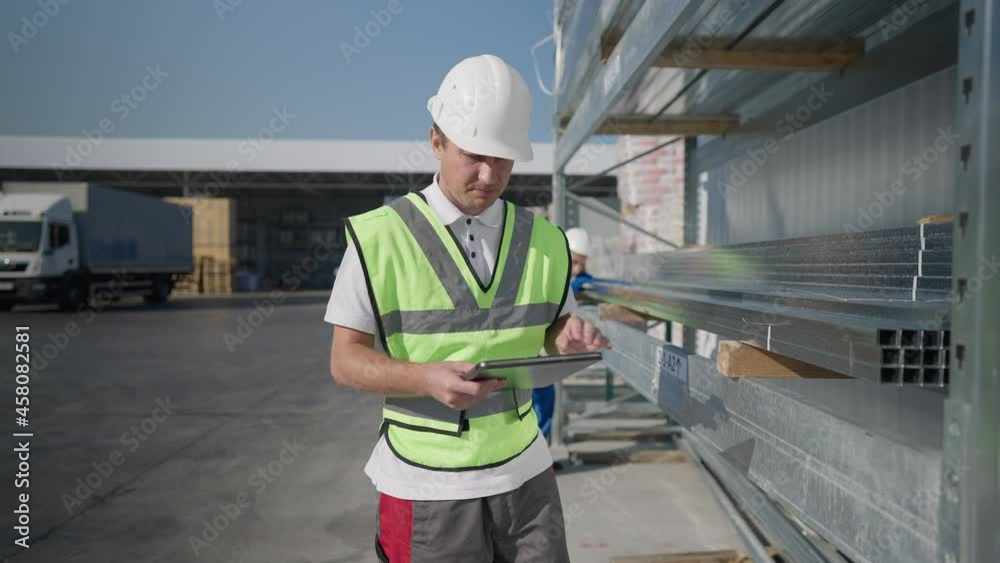 Confident serious Caucasian man in hard hat walking along row of delivered metal products at warehouse with blurred Middle Eastern employee checking quality visually at background. Slow motion Confident serious Caucasian man in hard hat walking along row of delivered metal products at warehouse with blurred Middle Eastern employee checking quality visually at background. Slow motion