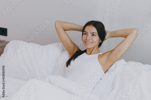 Marvelous happy young woman in white tank top sit in bed hands folded under head look aside rest relax spend time in bedroom lounge home in own room house wake up dream be lost in reverie good day