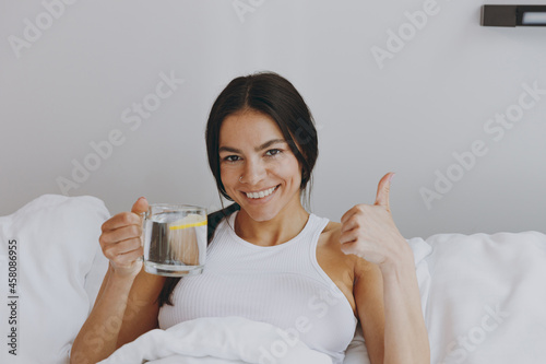 Happy young woman in white tank top sit in bed hold drink clear fresh water with lemon showing thumb up like gesture in bedroom lounge home in own room house wake up dream be lost in reverie good day.