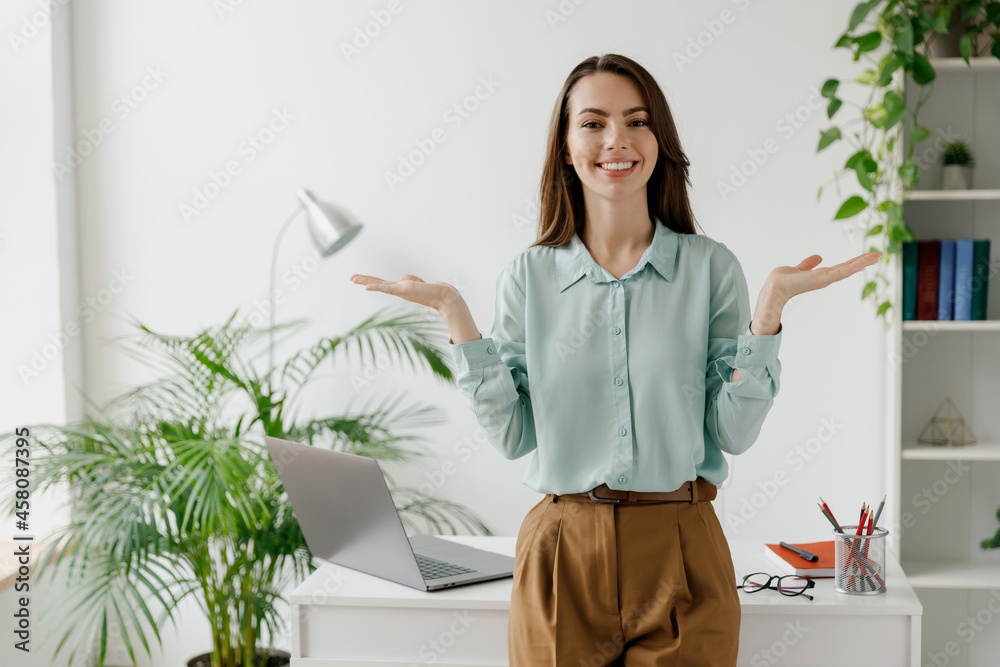 Young fun smiling successful employee business woman in casual blue shirt work look camera stand spread hands near workplace desk with laptop pc computer at office indoors. Achievement career concept. Young fun smiling successful employee business woman in casual blue shirt work look camera stand spread hands near workplace desk with laptop pc computer at office indoors. Achievement career concept.
