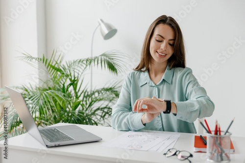 Young smiling happy successful employee business woman 20s in blue shirt look at smartwatch time sit work at workplace white desk with laptop pc computer at office indoors. Achievement career concept. photo