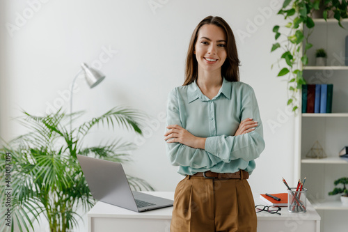Young smiling happy successful employee business woman in casual blue shirt work stand hold hands crossed folded at workplace desk with laptop pc computer at office indoors Achievement career concept