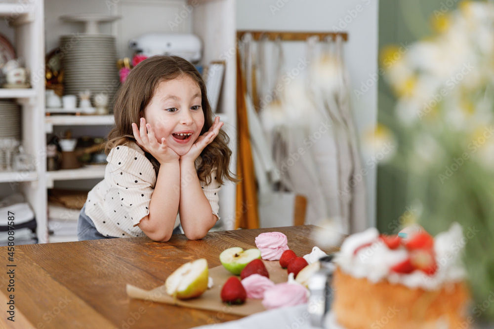 Young surprised fun happy positive little kid child girl daughter sitting at table near fruits for sweet pie cake, learn how to cook in green kitchen indoors. People lifestyle, Mother's Day concept Young surprised fun happy positive little kid child girl daughter sitting at table near fruits for sweet pie cake, learn how to cook in green kitchen indoors. People lifestyle, Mother's Day concept