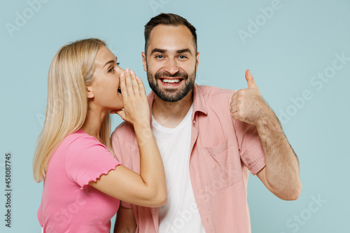 Young couple two friends family man woman in casual clothes whisper gossip tell secret behind her hand sharing news together show thumb up isolated on pastel plain light blue color background studio.