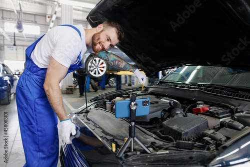 Young technician car mechanic man in blue overalls white t-shirt talk mobile cell on phone stand show troubleshooting process fix problem with raised hood work in vehicle repair shop workshop indoors