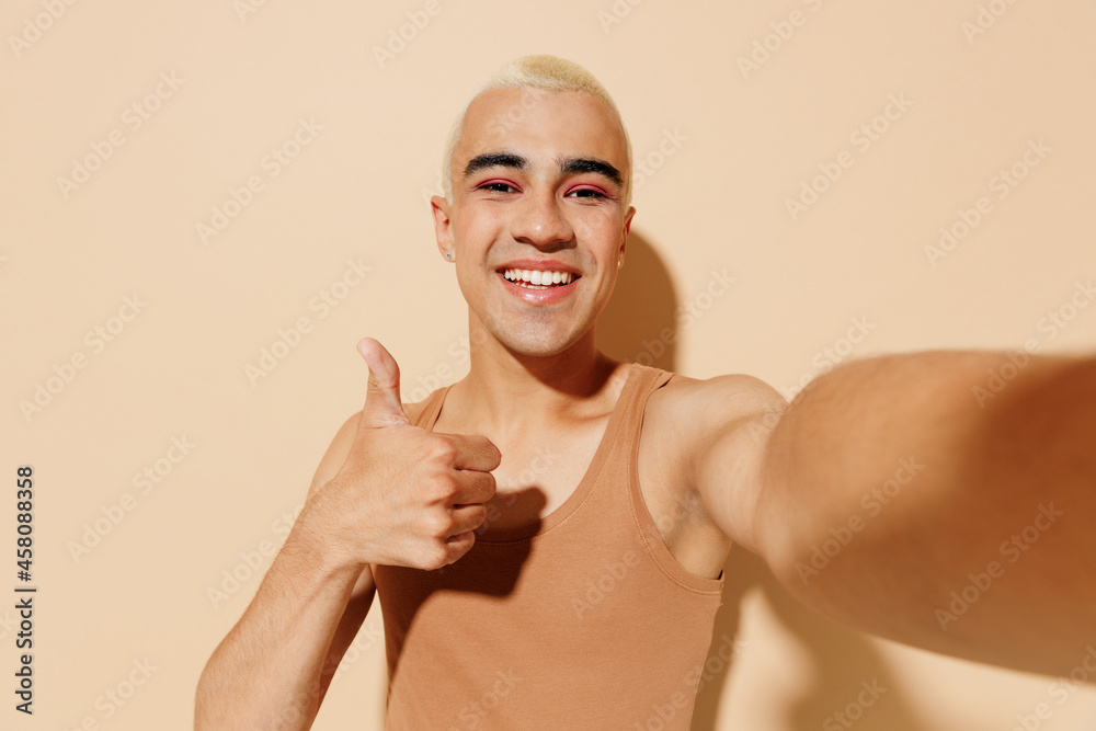 Close up young blond latin gay man with make up in beige tank shirt doing selfie shot pov on mobile phone show thumb up isolated on plain light ocher background studio People lgbt lifestyle concept. Close up young blond latin gay man with make up in beige tank shirt doing selfie shot pov on mobile phone show thumb up isolated on plain light ocher background studio People lgbt lifestyle concept.
