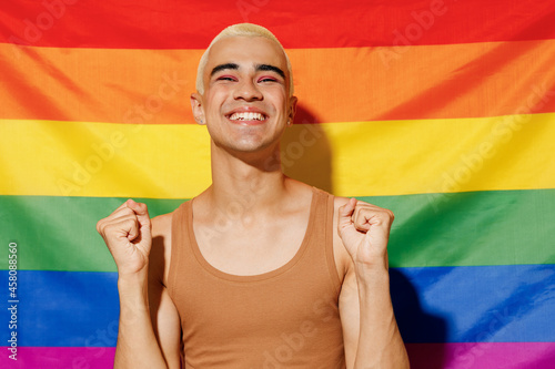 Young queer excited smiling satisfied happy blond latin gay man 20s with make up in beige tank shirt do winner gesture stand on rainbow flag background studio portrait People lgbt lifestyle concept.