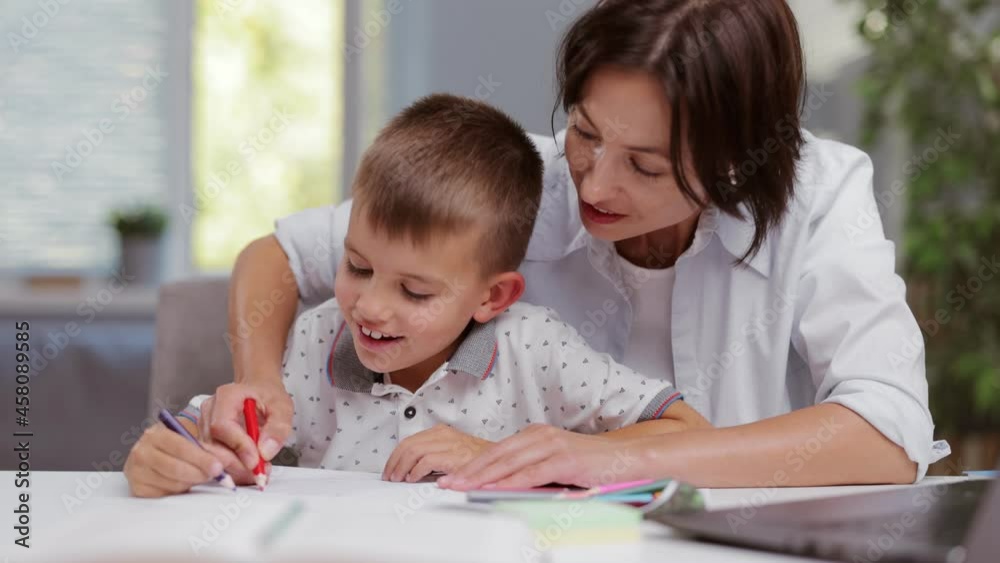 Positive boy and mature woman sitting together at desk and drawing with colorful pencils in album. Mother and son spending time at home. Education process.