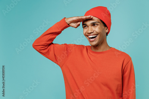 Young smiling fun cheerful cool happy african american man in red shirt hat hold hand at forehead look far away distance isolated on plain pastel light blue background studio People lifestyle concept