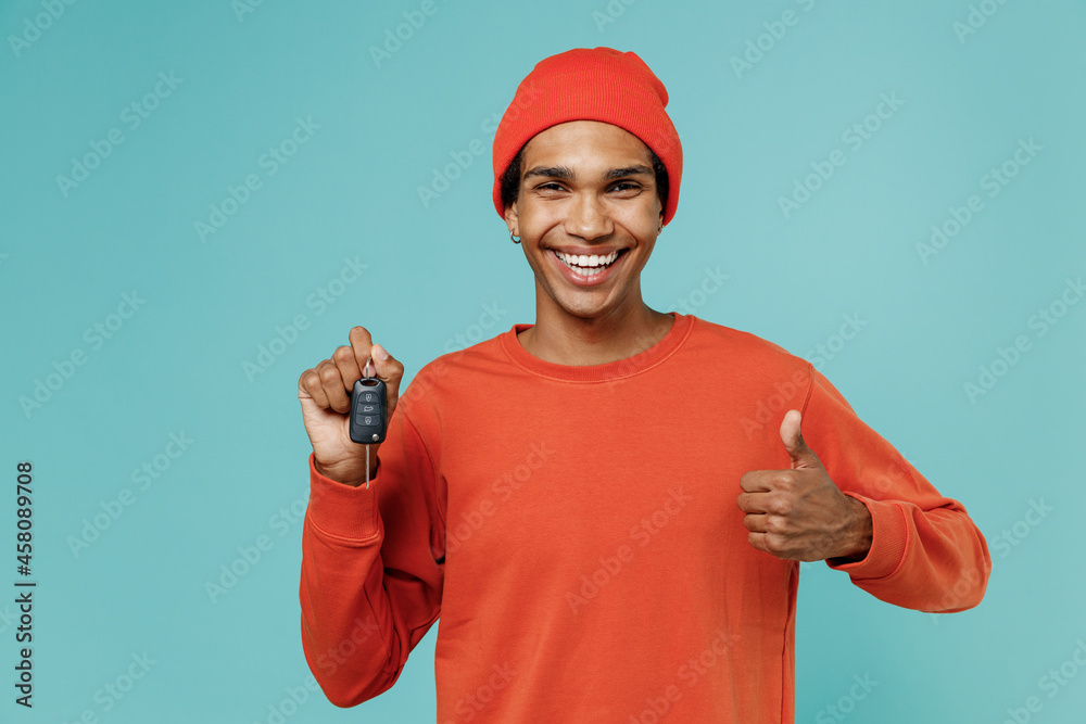 Young smiling fun excited happy african american man in orange shirt hat hold car key show thumb up gesture isolated on plain pastel light blue background studio portrait. People lifestyle concept
