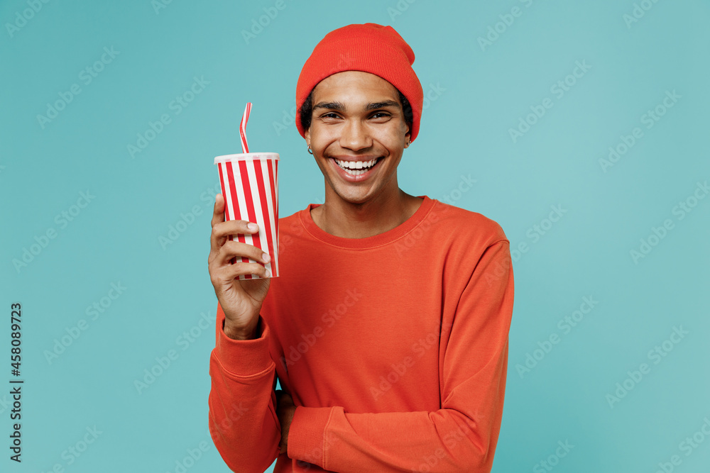 Young smiling satisfied happy african american man 20s in orange shirt hat hold plastic cup of soda cola water isolated on plain pastel light blue background studio portrait. People lifestyle concept. Young smiling satisfied happy african american man 20s in orange shirt hat hold plastic cup of soda cola water isolated on plain pastel light blue background studio portrait. People lifestyle concept.
