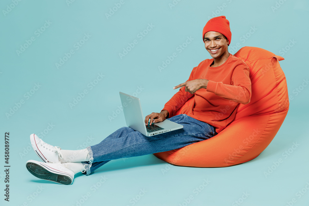 Full body young smiling satisfied happy african american man in orange  shirt hat sit in bag