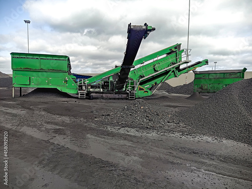 Mechanized loading of bulk cargo. Heaps of gravel on the dock and a harvester with transport belts. a loading harvester loads coal onto an offshore ship for delivery