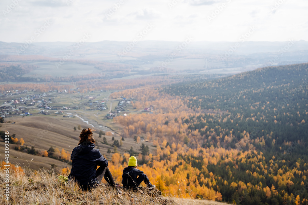 A woman in dark clothes and a boy in a bright yellow hat sit on the mountain, looking forward at the forest. Part of the forest managed to turn yellow.