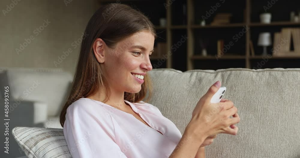 Side view of gen z teenage female satisfied user of modern mobile phone reclining on couch browsing internet downloading software. Young lady surfing social networks on cell choose goods at web store