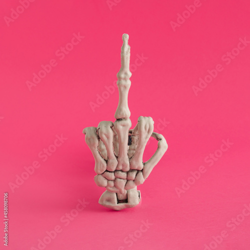 Halloween arrangement made of a skeleton hand that shows the middle finger. Trendy pink background. Minimal scary concept. Halloween party inspiration.