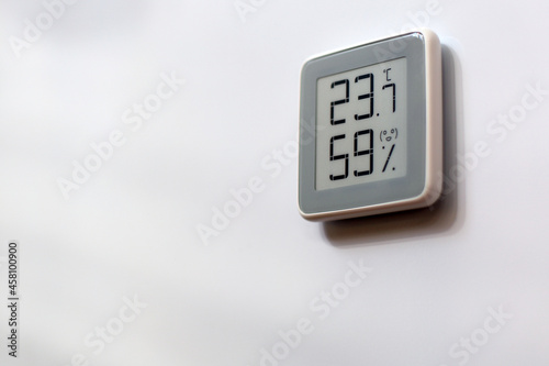 Electronic indoor gray plastic digital battery powered thermometer on white wall showing temperature, humidity level in house, smiling smiley face indoors in daylight sun