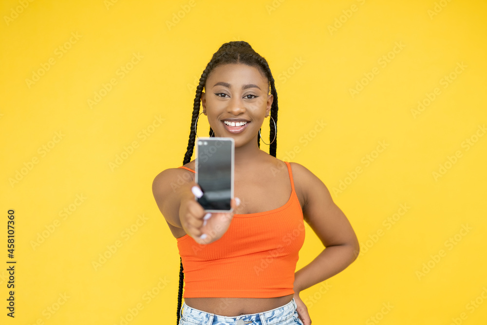 Phone app. Mobile ad. Gadget service. Cheerful smiling African woman recommending defocused cell screen mockup isolated on yellow background. Phone app. Mobile ad. Gadget service. Cheerful smiling African woman recommending defocused cell screen mockup isolated on yellow background.