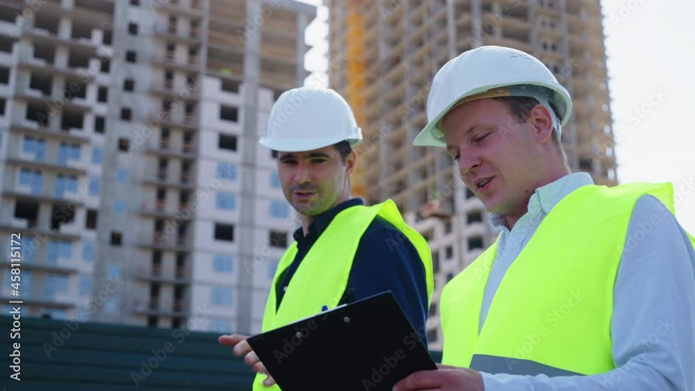 Men wearing hard hats and safety vests walking at construction site, one holding map case. Tracking shot architect and contractor checking plans or blueprints of multistory buildings Men wearing hard hats and safety vests walking at construction site, one holding map case. Tracking shot architect and contractor checking plans or blueprints of multistory buildings