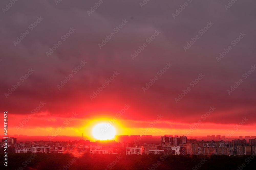 Hot red sun behind the silhouettes of city buildings. Summer sunset in the sky haze with night houses